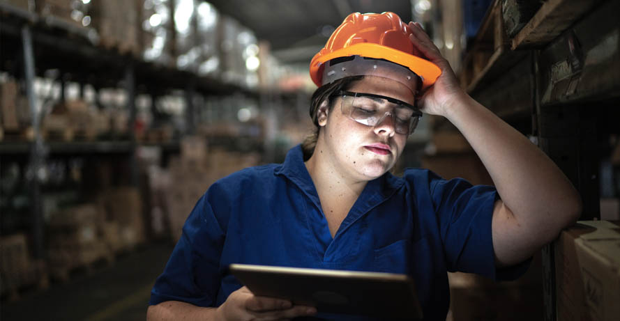 A woman in a warehouse wearing a hard hat. She struggles to say awake while doing her job