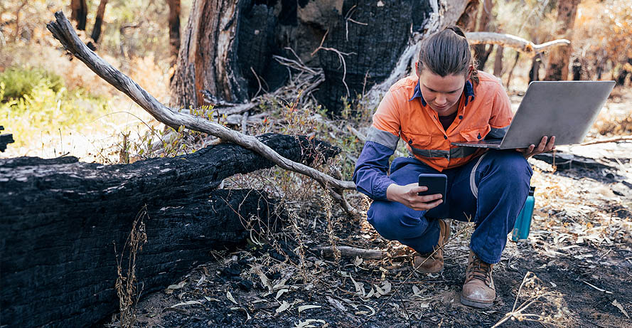 A woman gathering data with a laptop and cell phone after a forest fire