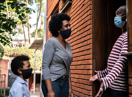 a woman and a boy greet an older woman standing in the doorway; all are wearing masks.