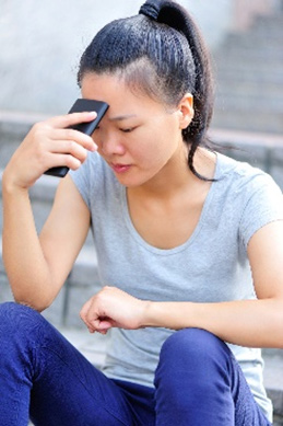 A young Asian woman sits on stairs outside. She is wearing blue pants, a grey t-shirt, and has her hair pulled back in a ponytail. Her left forearm is resting on her left knee and her right elbow rests on her right knee.  She holds her cell phone in her right hand and rests the top of the phone against her forehead. She appears to be deep in thought and perhaps sad..