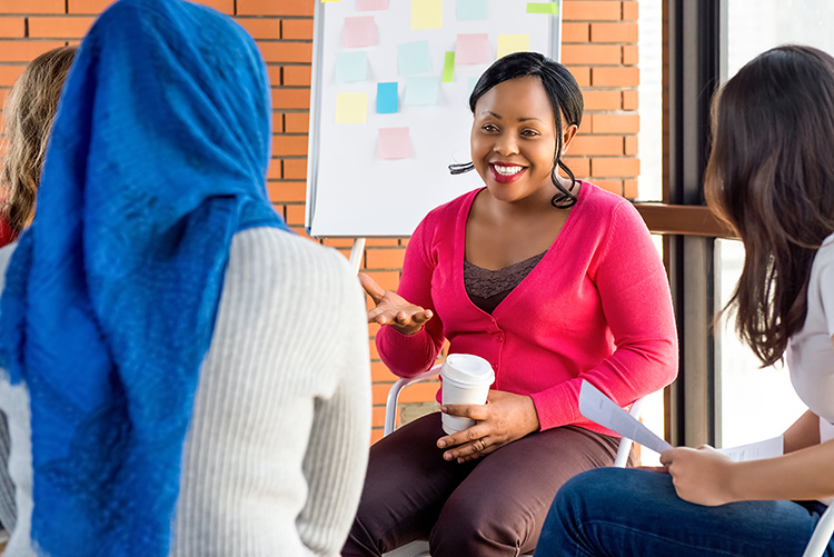 A group of public health planners and mental health professionals meet to build partnerships prior to an emergency. A group of four women sits in a circle talking. The woman in focus at the center of the picture is wearing a pink sweater, brown pants, and is holding a cup of coffee. She has a big smile on her face, and she is using her hands expressively as she talks.
