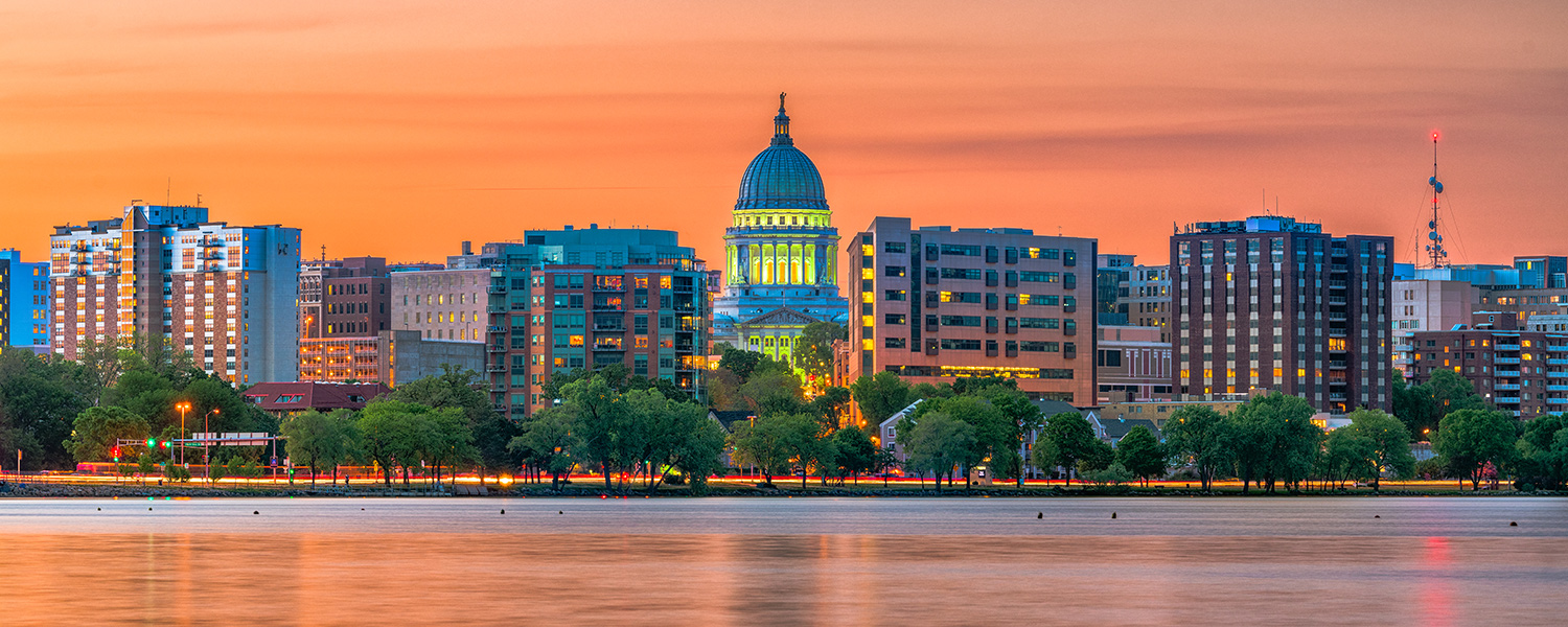 Landscape picture of a city along a river at sunset. In the foreground, the river runs left to right with trees lining the bank on the far side. In the center of the photo is the city’s government building all lit up. Other city buildings fill in the photo on both the right and left of the government building.