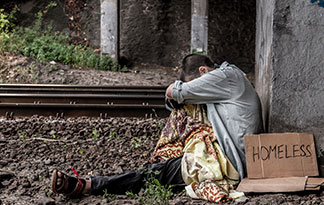 Person sitting next to railroad tracks with a cardboard sign that says, 'Homeless'