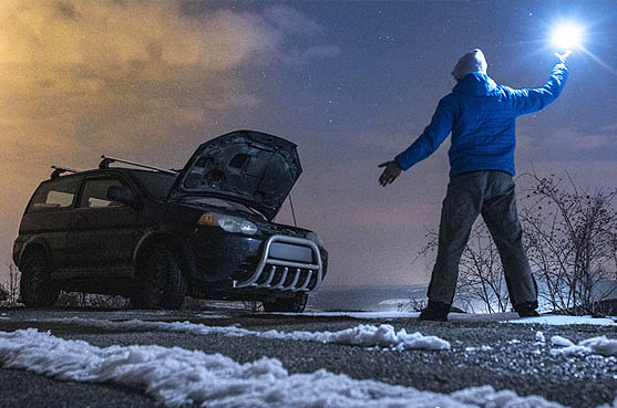 A person holding up a flashlight. Their car is broken down on the side of a snowy road.